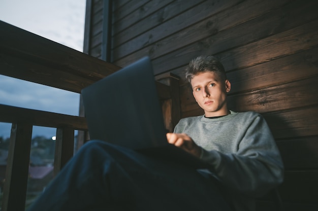 Giovane uomo seduto su una sedia su un balcone utilizzando un computer portatile