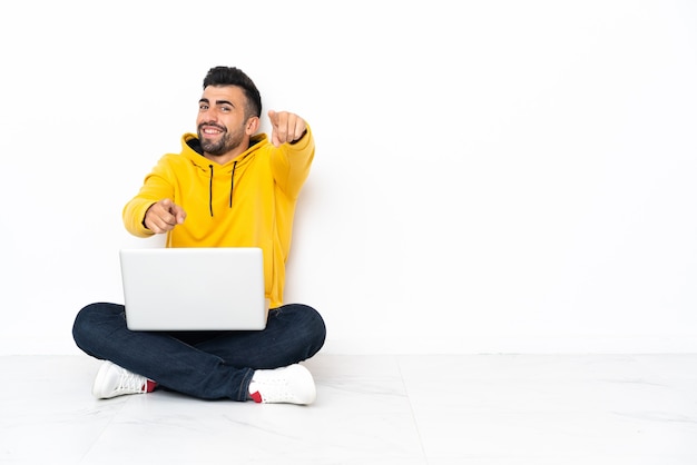 Giovane uomo seduto sul pavimento con il suo laptop punta il dito contro di te mentre sorride