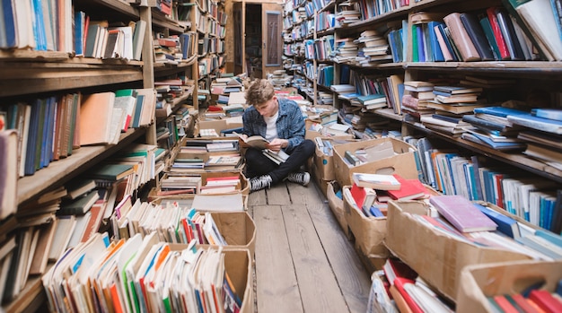 Giovane uomo seduto sul pavimento in una vecchia biblioteca pubblica e libri di lettura.