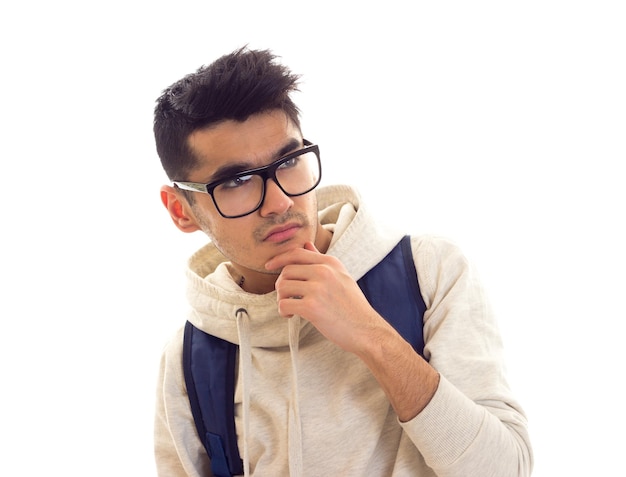 Giovane uomo senza allegria con capelli neri in felpa bianca con occhiali neri e zaino blu in studio