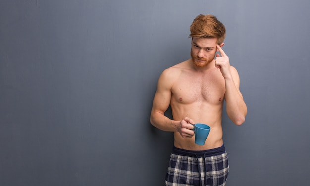 Giovane uomo senza camicia rossa pensando a un&#39;idea. Tiene una tazza da caffè.