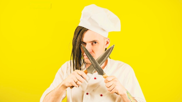 Giovane uomo serio vestito da chef con coltelli su sfondo giallo Cuoco maschio con cappello bianco e camicia con coltelli da cucina incrociati