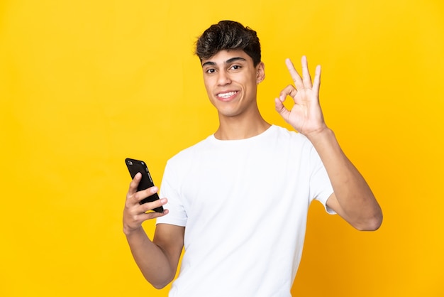Giovane uomo sopra la parete gialla isolata utilizzando il telefono cellulare e facendo segno OK