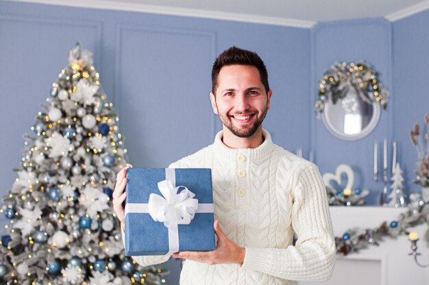 Giovane uomo sorridente attraente in posa in studio splendidamente decorato per Natale con scatola regalo quadrata blu con nastro bianco Concetto di vacanze invernali