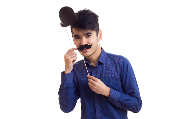 Giovane uomo sorridente con i capelli scuri in camicia blu che tiene bastoncini di cartone di baffi e cappello nero su sfondo bianco in studio