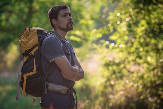 Giovane uomo sorridente dello zaino in natura della foresta di estate. Studente adulto maschio bello felice che esamina macchina fotografica che cammina facendo un'escursione nel fondo della foresta. Borsa da scuola o concetto di viaggio zaino in spalla.