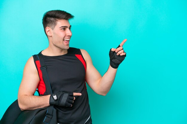 Giovane uomo sportivo caucasico con borsa sportiva isolata su sfondo blu che punta il dito di lato e presenta un prodotto