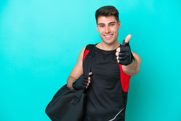 Giovane uomo sportivo caucasico con borsa sportiva isolata su sfondo blu con i pollici in su perché è successo qualcosa di buono