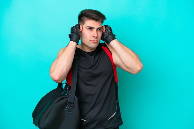 Giovane uomo sportivo caucasico con borsa sportiva isolata su sfondo blu frustrato e che copre le orecchie