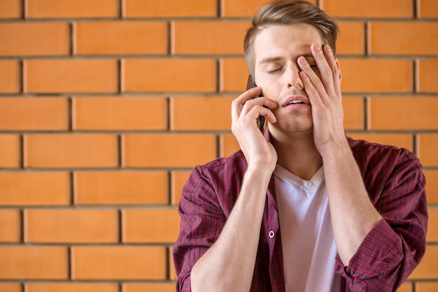 Giovane uomo stanco che parla sul telefono sul muro di mattoni