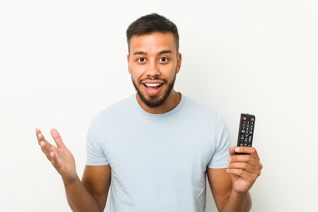 Giovane uomo sud-asiatico che tiene un controller tv che riceve una piacevole sorpresa, eccitato e alzando le mani.