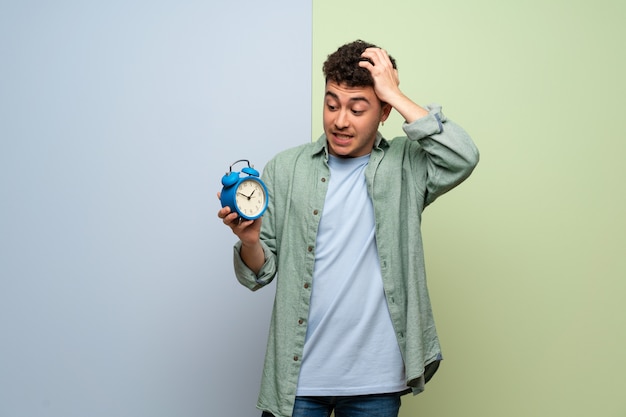 Giovane uomo sul muro blu e verde irrequieto perché è diventato tardi e in possesso di sveglia vintage