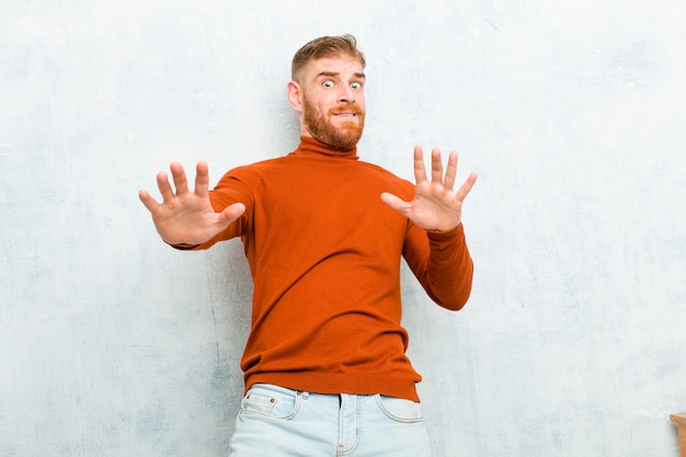 Giovane uomo testa rossa che indossa il collo di tartaruga sensazione di stupore e paura, temendo qualcosa di spaventoso, con le mani aperte sul davanti dicendo di stare lontano sul muro di cemento