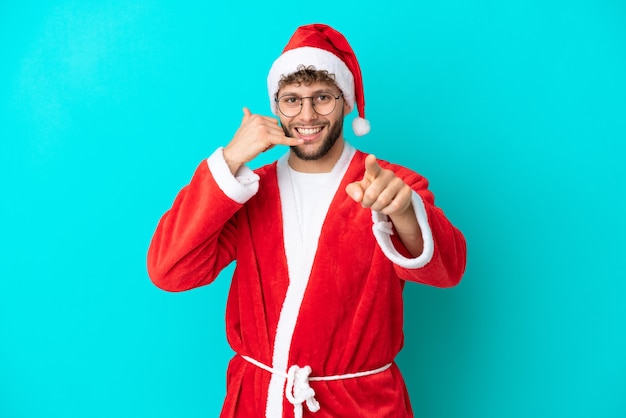 Giovane uomo travestito da Babbo Natale isolato su sfondo blu che fa il gesto del telefono e indica la parte anteriore