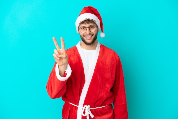 Giovane uomo travestito da Babbo Natale isolato su sfondo blu che sorride e mostra il segno della vittoria