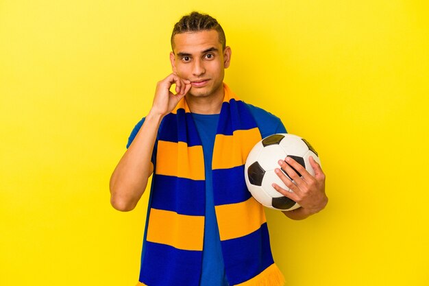 Giovane uomo venezuelano che guarda il calcio isolato su sfondo giallo con le dita sulle labbra mantenendo un segreto.