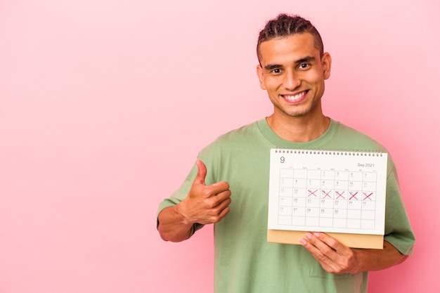 Giovane uomo venezuelano che tiene un calendario isolato su sfondo rosa sorridente e alzando il pollice thumb