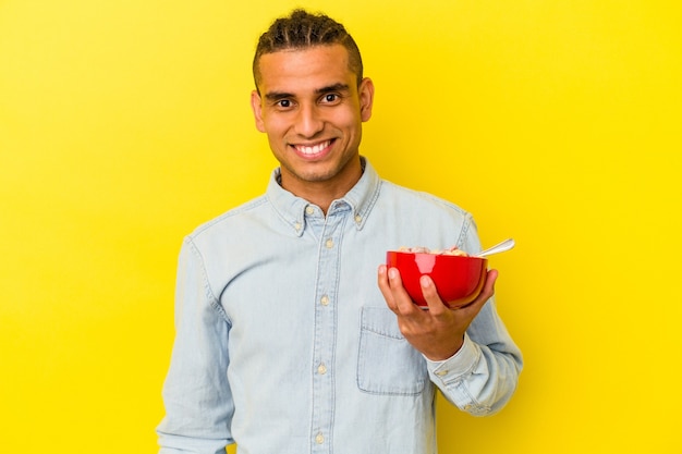 Giovane uomo venezuelano che tiene una ciotola di cereali isolata su sfondo giallo felice, sorridente e allegro.