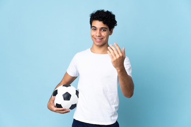 Giovane uomo venezuelano isolato sull'azzurro con pallone da calcio e facendo gesto in arrivo