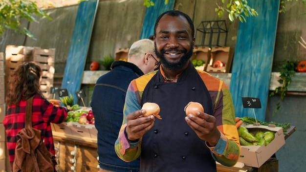 Giovane venditore maschio che presenta prodotti biologici naturali e parla con i clienti al mercato degli agricoltori. Mercato all'aperto con frutta e verdura biologica coltivata localmente, titolare della bancarella del mercato.