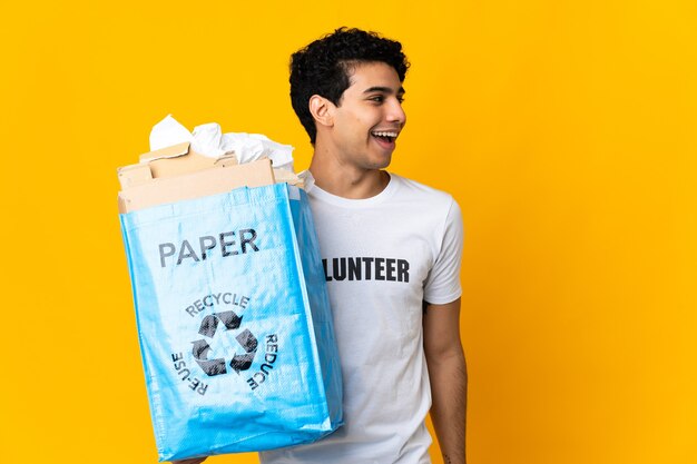 Giovane venezuelano che tiene un sacchetto di riciclaggio pieno di carta per riciclare ridendo in posizione laterale