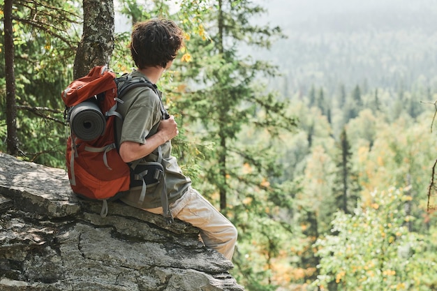 Giovane viaggiatore con zaino seduto su pietra in montagna e guardando la foresta