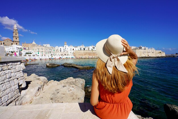 Giovane viaggiatore donna in abito arancione e cappello seduto sulla banchina del porto di Monopoli guardando la città storica dal mare Destinazione di viaggio in Puglia Italia