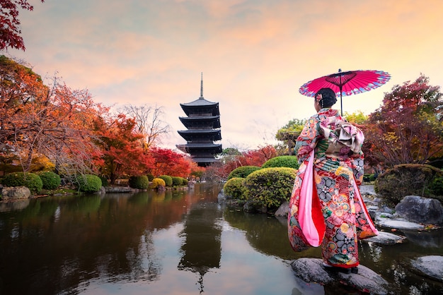 Giovane viaggiatore giapponese della ragazza in vestito tradizionale da kimino che sta in tempio di Toji con la pagoda di legno e la foglia di acero rossa nella stagione di autunno a Kyoto, Giappone.