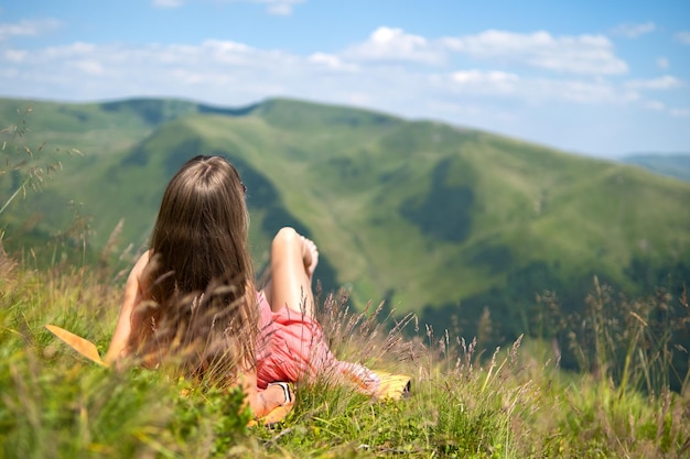 Giovane viaggiatrice felice in abito rosso che riposa sul pendio erboso verde in una giornata ventosa in montagne estive godendo della vista della natura.