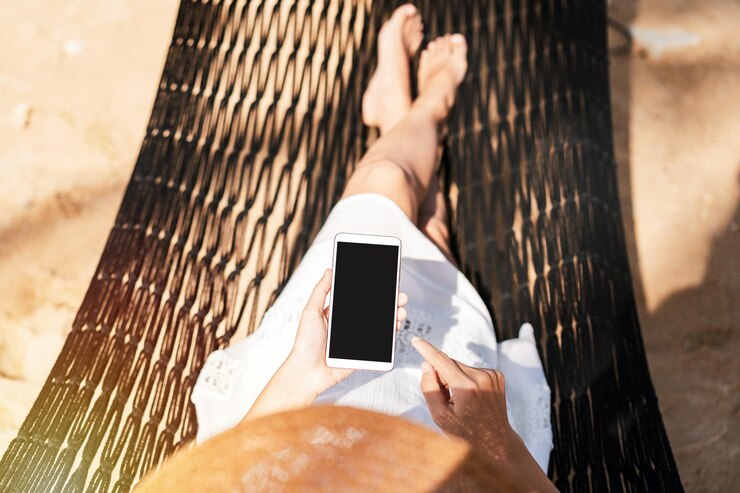 Giovane viaggiatrice sdraiata su un'amaca e che usa lo smartphone in spiaggia mentre viaggia per le vacanze estive