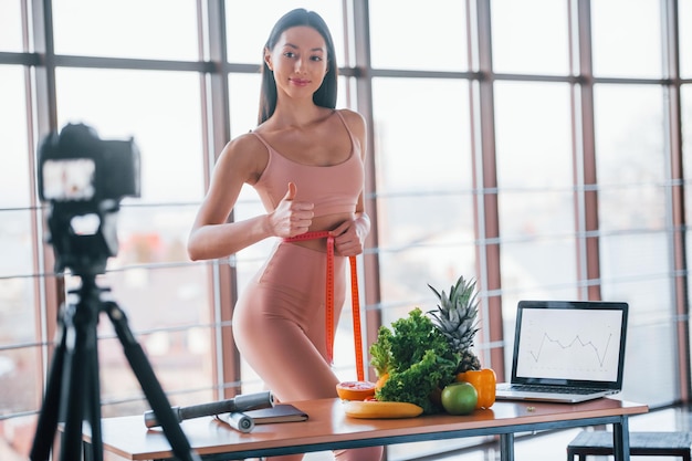 Giovane vlogger di fitness che fa video al chiuso stando vicino al tavolo con cibo sano e laptop.