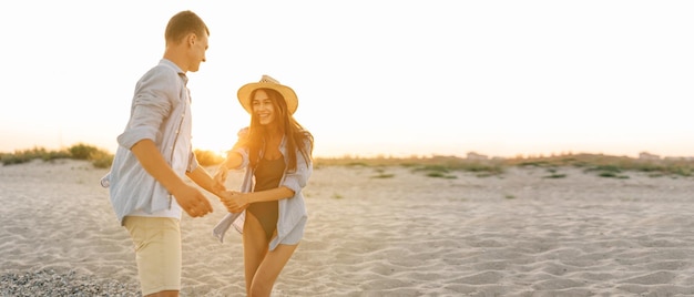 Giovani amanti felici uomo e donna che si tengono per mano camminano sulla spiaggia la sera durante una vacanza in un resort tropicale Banner