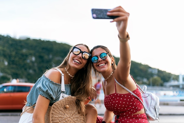 Giovani amiche felici in occhiali da sole seduti e facendo un selfie sul telefono cellulare della strada della città