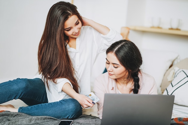 Giovani amici di ragazze spensierate del brunette nell'acquisto casuale online dalla carta di credito sul computer portatile, acquistante dalla casa