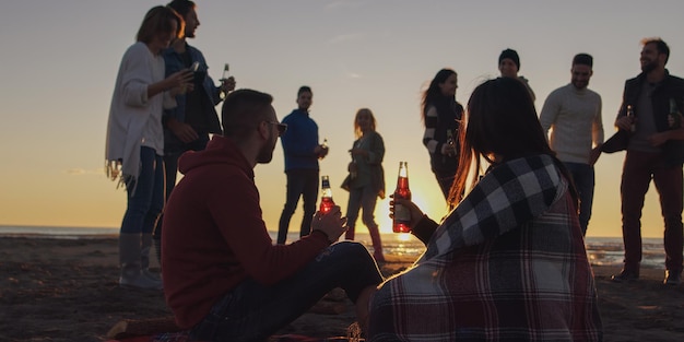 Giovani amici spensierati felici che si divertono e bevono birra vicino al fuoco delle ossa sulla spiaggia mentre il sole inizia a tramontare