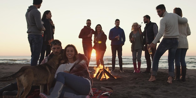 Giovani amici spensierati felici che si divertono e bevono birra vicino al fuoco delle ossa sulla spiaggia mentre il sole inizia a tramontare