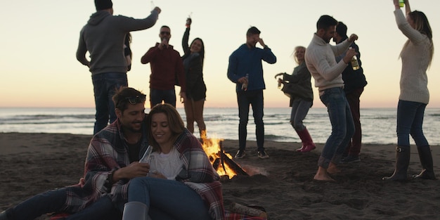 Giovani amici spensierati felici che si divertono e bevono birra vicino al fuoco delle ossa sulla spiaggia mentre il sole inizia a tramontare