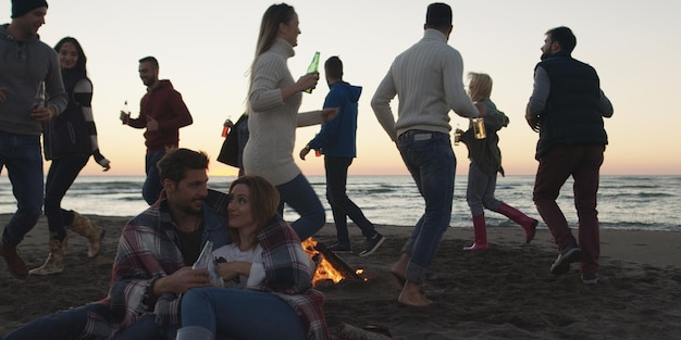 Giovani amici spensierati felici che si divertono e bevono birra vicino al fuoco delle ossa sulla spiaggia mentre il sole inizia a tramontare