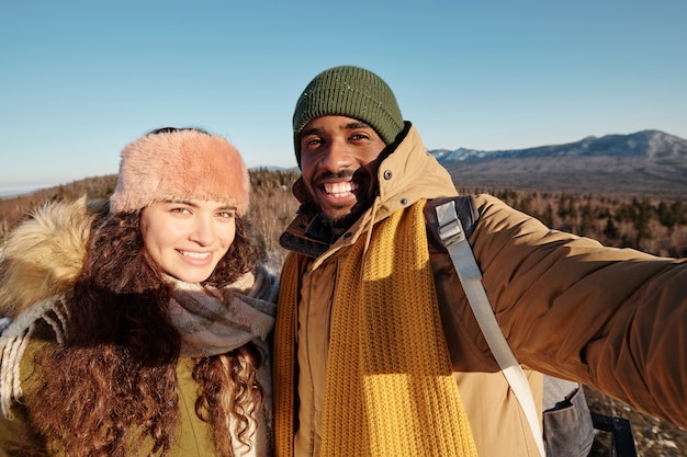 Giovani appuntamenti allegri in abbigliamento invernale che si fanno selfie davanti alla telecamera contro il cielo blu e le montagne in una giornata di sole mentre si rilassano nel tempo libero