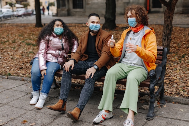 Giovani attraenti in maschere mediche che si rilassano insieme all'aperto. Amici multirazziali che godono del tempo libero seduti sulla panca di legno al parco d'autunno.