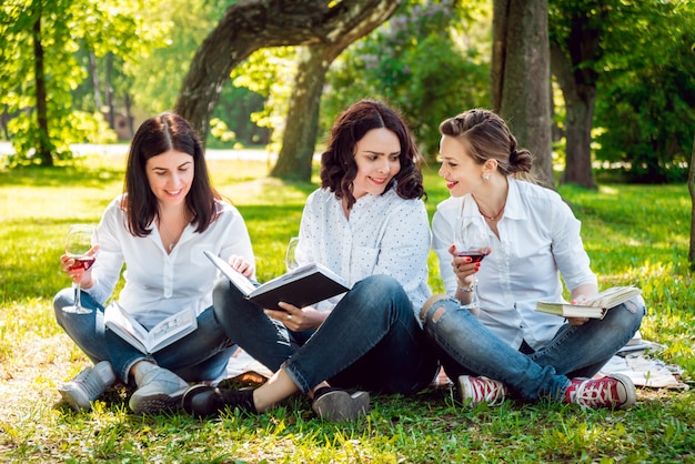 Giovani belle ragazze con bicchiere di vino rosso e libri nel parco.