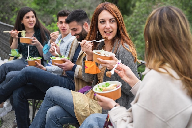 Giovani che si radunano al parco. Riunione dei migliori amici che mangiano insieme insalate in ciotole di cartone da asporto seduti all'aperto e divertirsi.