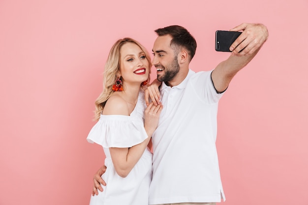 Giovani coppie allegre attraenti che stanno insieme isolate sopra il rosa, prendendo un selfie