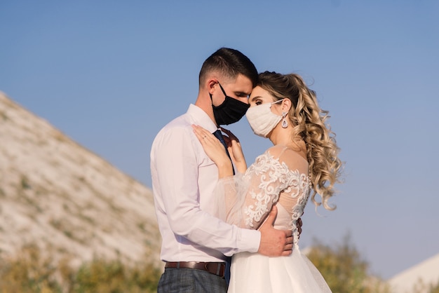 Giovani coppie amorose che camminano in maschere mediche nel parco durante la quarantena il giorno delle nozze.