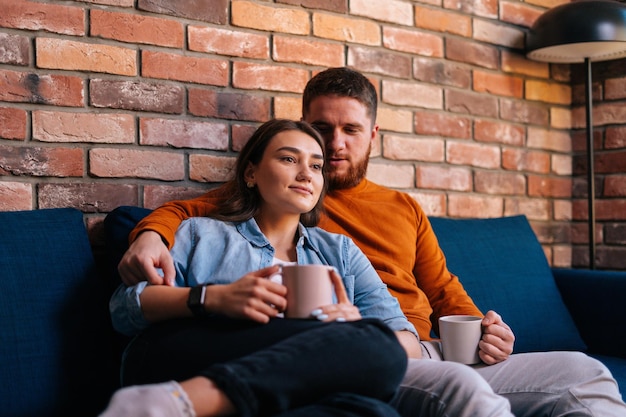 Giovani coppie amorose che si abbracciano, tengono in mano tazze di bevanda calda e guardano film romantici in TV la sera insieme seduti su un comodo divano nell'accogliente soggiorno. Concetto di vita domestica.