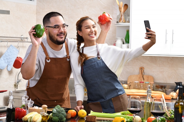 Giovani coppie asiatiche amorose che cucinano nella cucina che fanno insieme il cibo sano divertendosi e usando lo smartphone per prendere i selfie