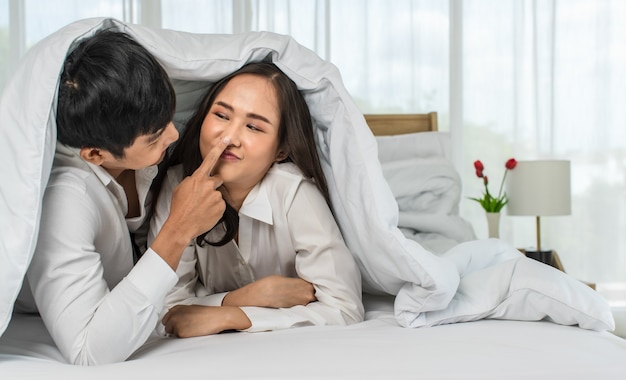 Giovani coppie asiatiche attraenti felici che si prendono in giro a letto con il fondo bianco della tenda.