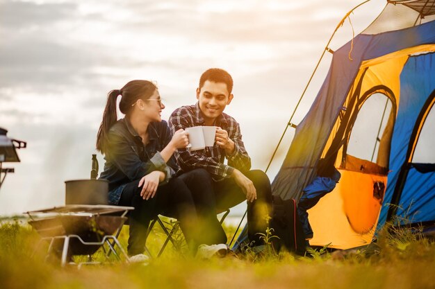 Giovani coppie asiatiche felici che si siedono sulla sedia di picnic che beve tè e caffè mentre si accampano in tenda in riva al lago