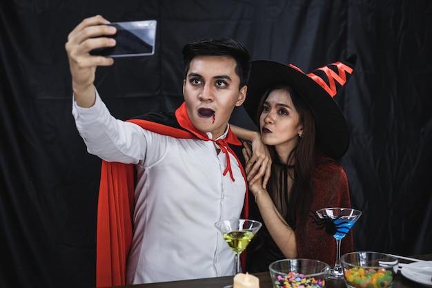 Giovani coppie asiatiche in costume da strega e dracula con selfie da cellulare durante la festa di Halloween. Le coppie in costume celebrano il fondo del panno nero della festa di Halloween.