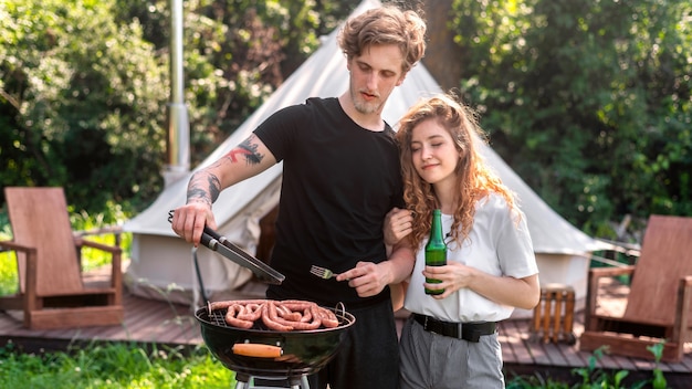 Giovani coppie che friggono carne alla griglia e bevono birra. Verde intorno. Glamping